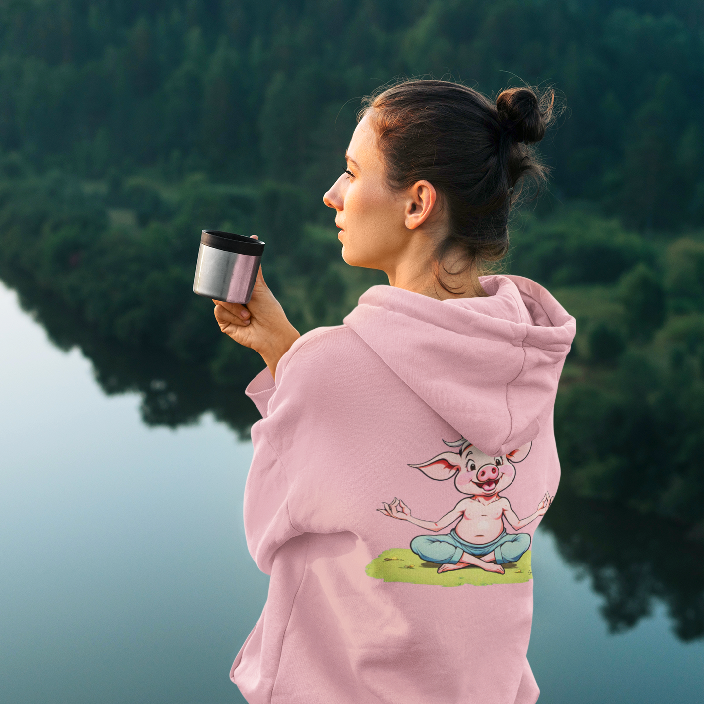 Eine Frau mit einem rosafarbenen Hoodie mit einem Schweinchen im Comic-Stil, das Yoga macht auf dem Rücken, schaut auf einen See.