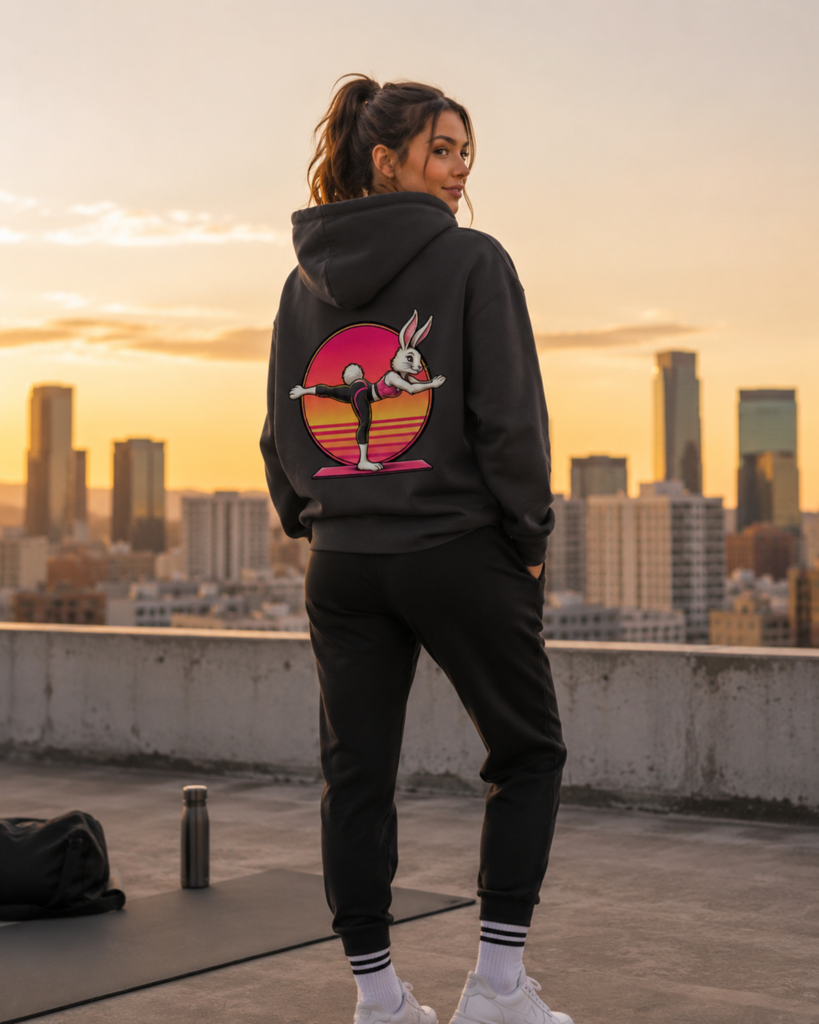 Eine junge hübsche Frau steht vor einer Skyline und trägt einen Hoodie von sporty animals in der Farbe Asphalt. Auf dem Rücken des Hoodies ist das Motiv eines süßen Hasen im Comic-Stil, der Pilates macht.