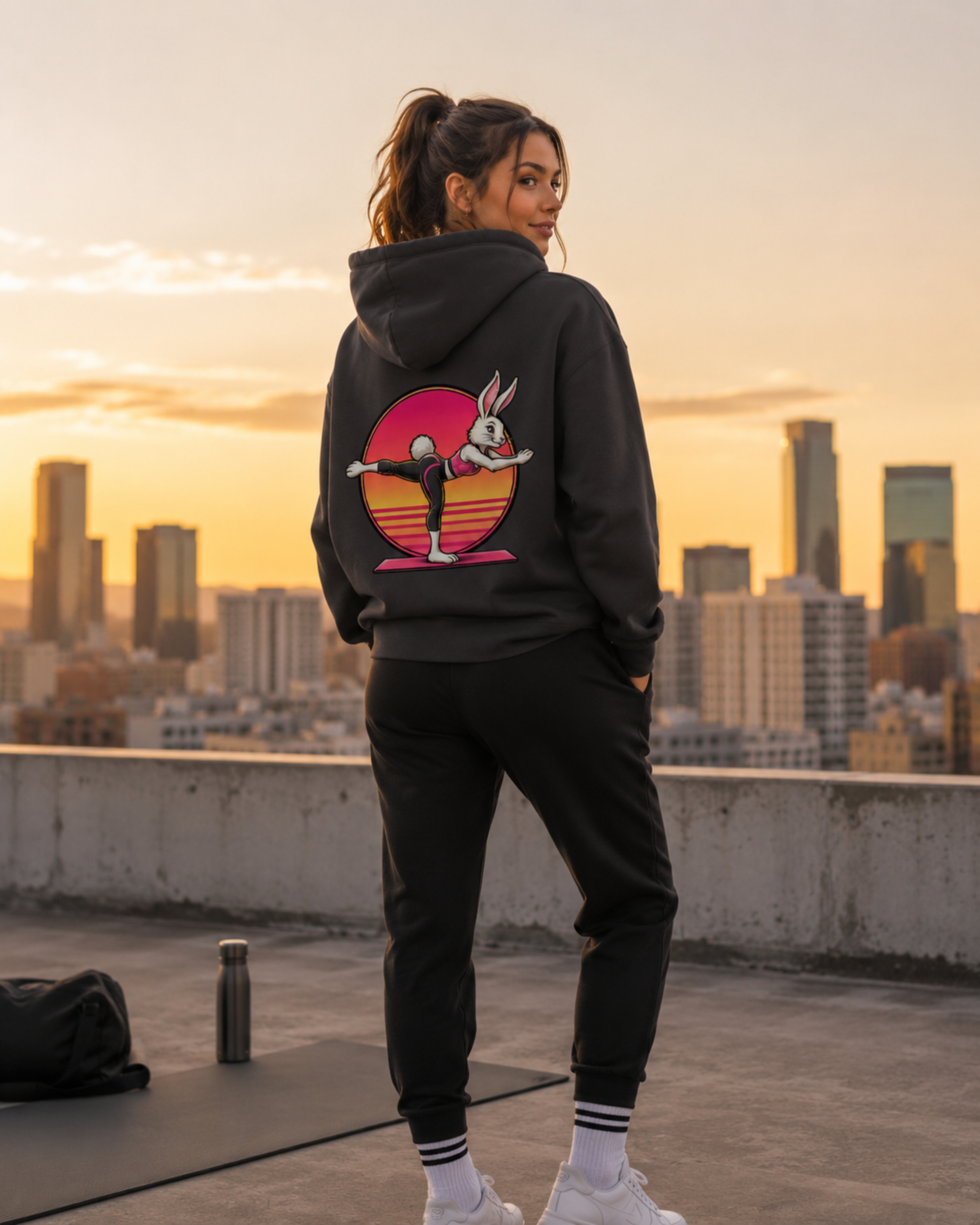Eine junge hübsche Frau steht vor einer Skyline und trägt einen Hoodie von sporty animals in der Farbe Asphalt. Auf dem Rücken des Hoodies ist das Motiv eines süßen Hasen im Comic-Stil, der Pilates macht.
