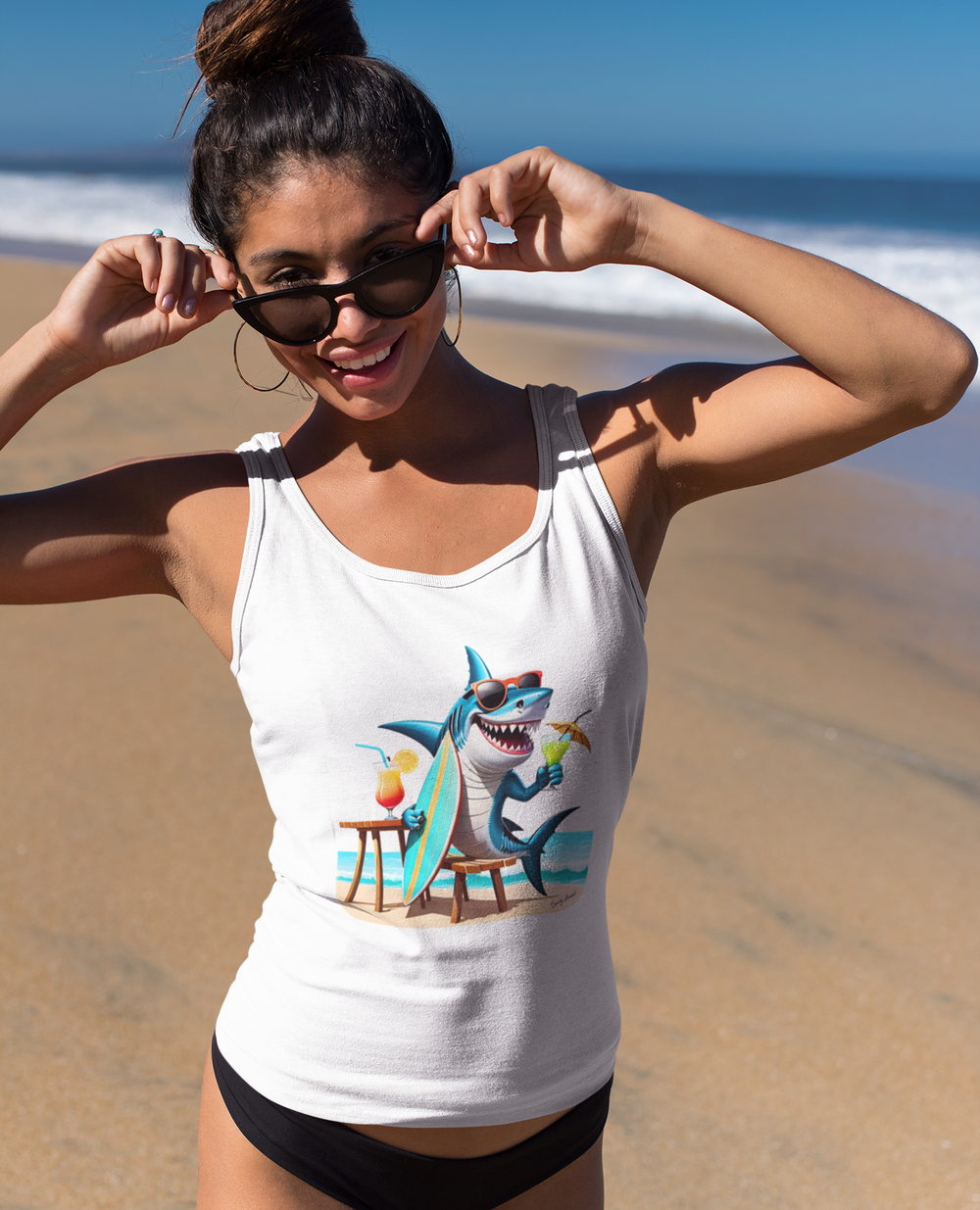 Frau am Strand trägt ein weißes Tank Top. Darauf abgebildet ist ein lustiger Hai im Comic-Stil der am Strand sitzt und ein Surfbrett in der Flosse hält
